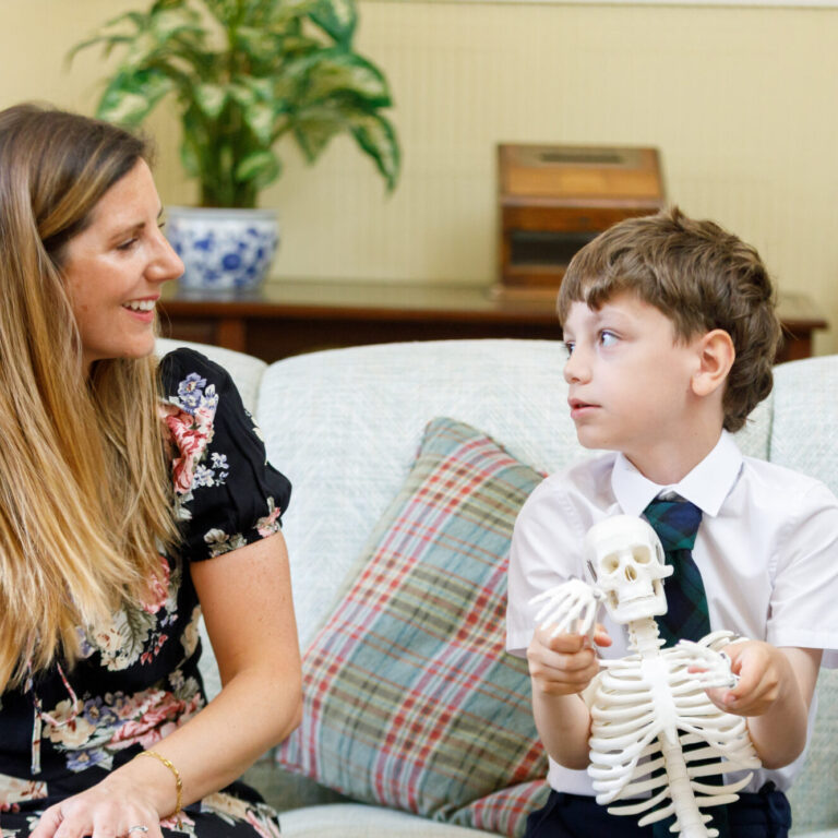 Young boy sitting with teacher and explaining about the skeleton he is holding.